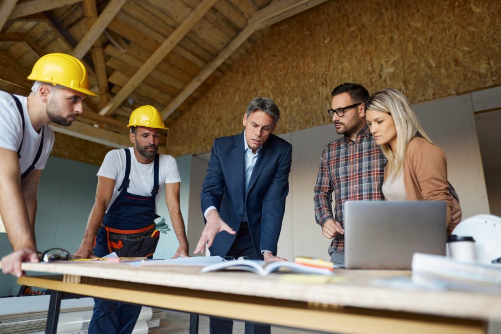Architect and manual workers cooperating with young couple while analyzing rebuilding plans at construction site.